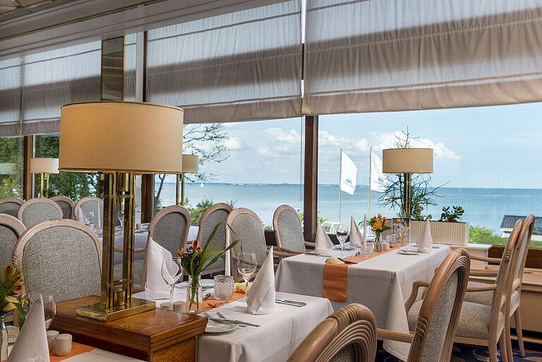 Elegant restaurant with set tables and a sea view, decorated with lamps and flowers for a fine dining experience.