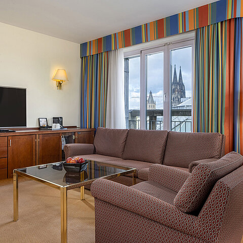 Living room of Senior Suite at Maritim Hotel Köln with sofa, TV, and Cologne Cathedral view through the windows.