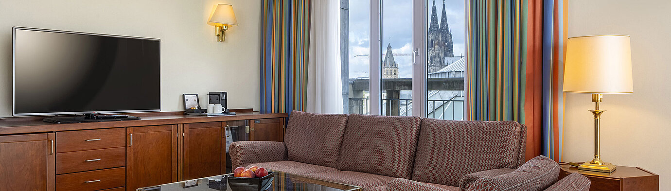 Living room of Senior Suite at Maritim Hotel Köln with sofa, TV, and Cologne Cathedral view through the windows.