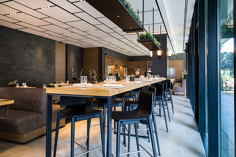 Bright restaurant with high wooden tables, modern chairs and hanging plants at Maritim Hotel Plaza Tirana