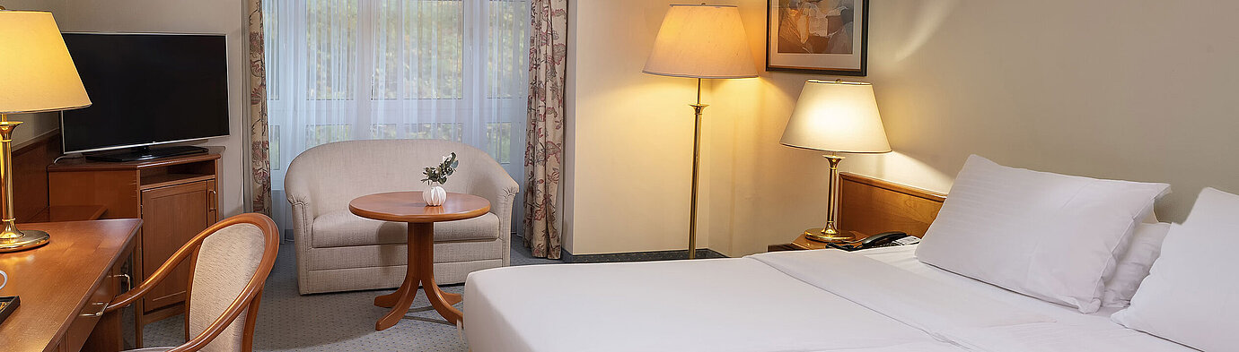 Classic room with double bed, desk, TV, seating area and large window in a bright interior design.