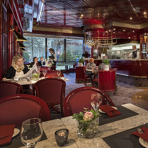 The image shows the cozy "Brasserie" café at the Maritim Hotel Bonn with red decor and a relaxed atmosphere.