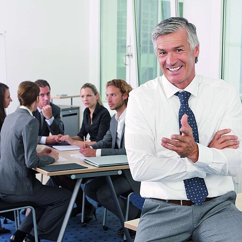 Person in the foreground smiling while a business team works together at a meeting table in the background.