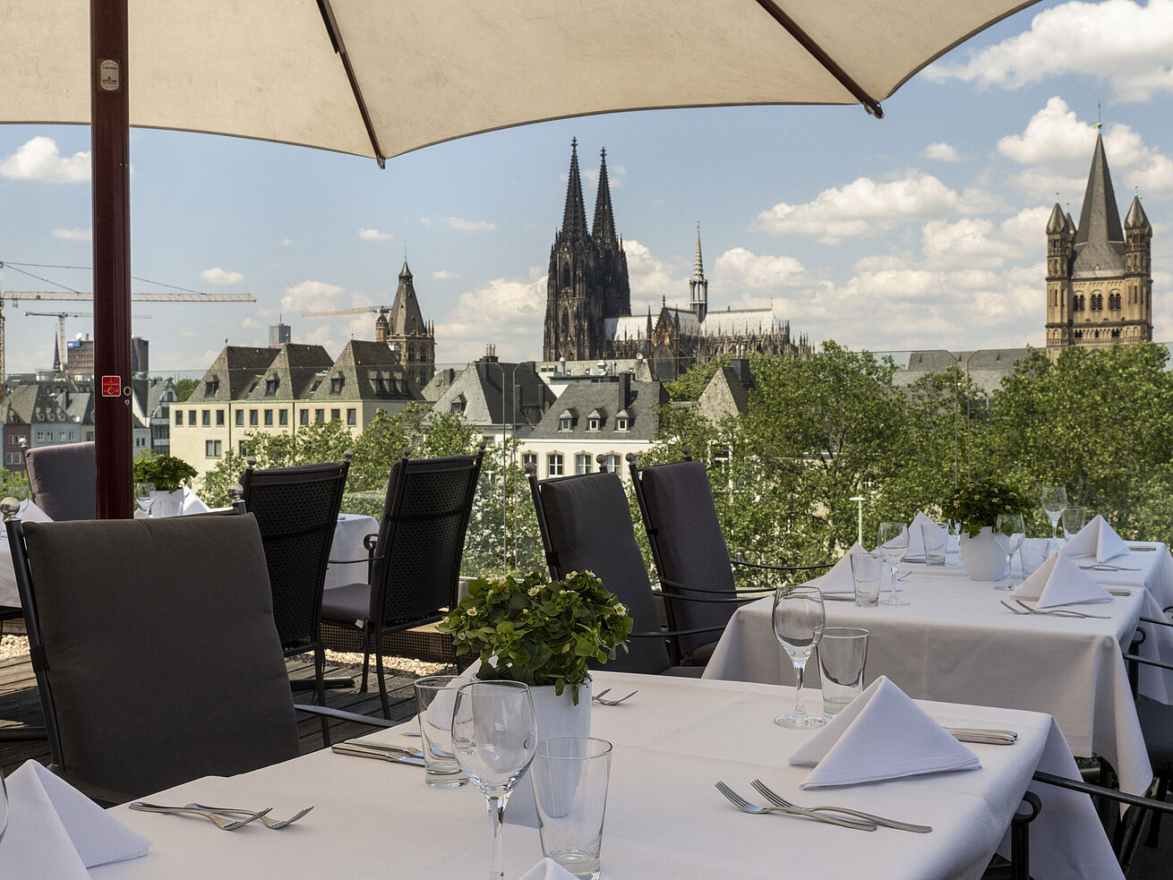 Rooftop terrace of Maritim Hotel Cologne overlooking the Rhine and Hohenzollern Bridge, with set tables under umbrellas.