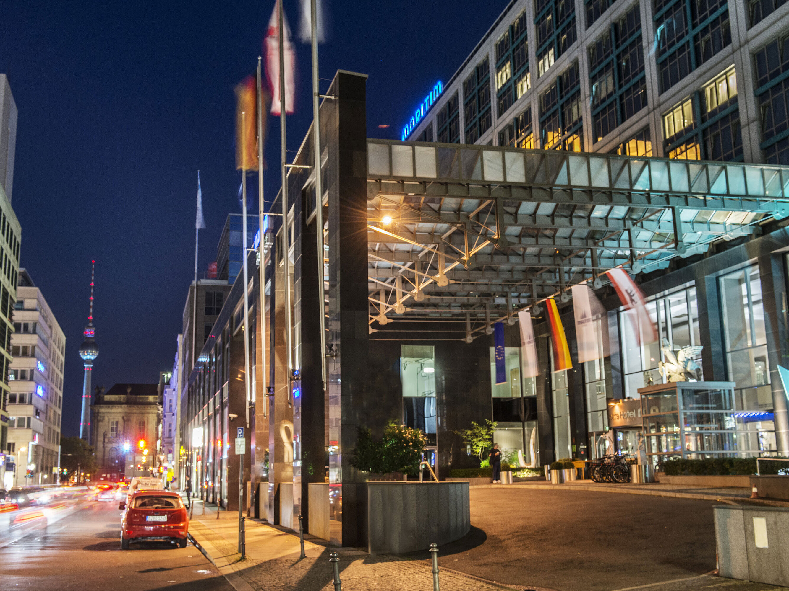 Night view of the illuminated entrance of Maritim proArte Hotel Berlin with TV tower in background and flags.