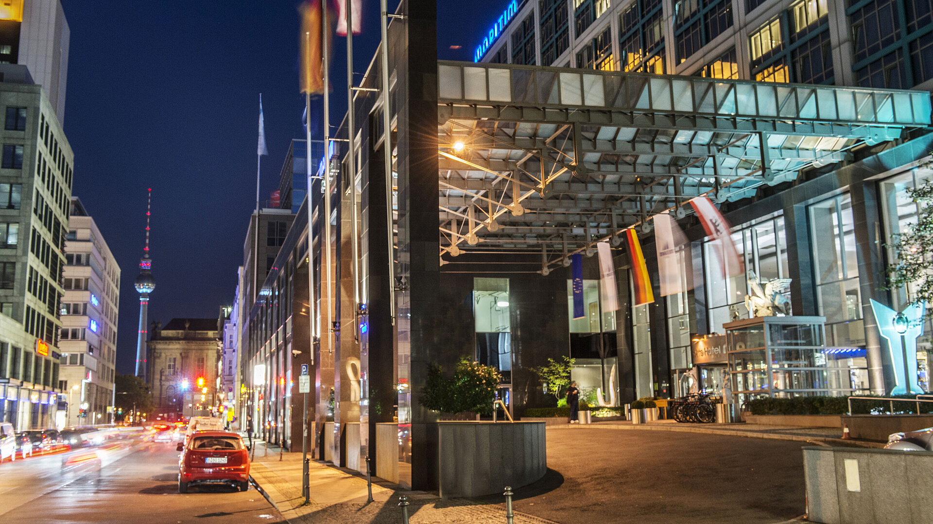 Night view of the illuminated entrance of Maritim proArte Hotel Berlin with TV tower in background and flags.
