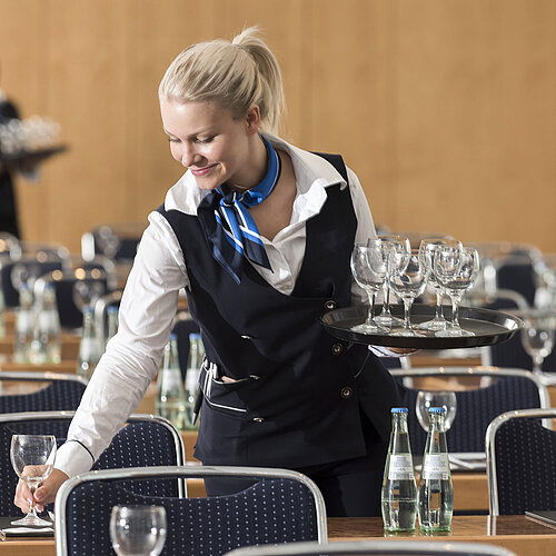 A service staff member at the Maritim Berlin sets up a conference room, carrying a tray of glasses and placing water bottles.