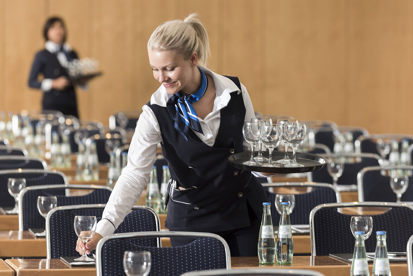 A service staff member at the Maritim Berlin sets up a conference room, carrying a tray of glasses and placing water bottles.