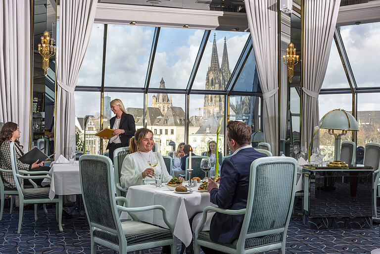 Restaurant "Bellevue" at Maritim Hotel Cologne with guests dining and a view of Cologne Cathedral through large windows.