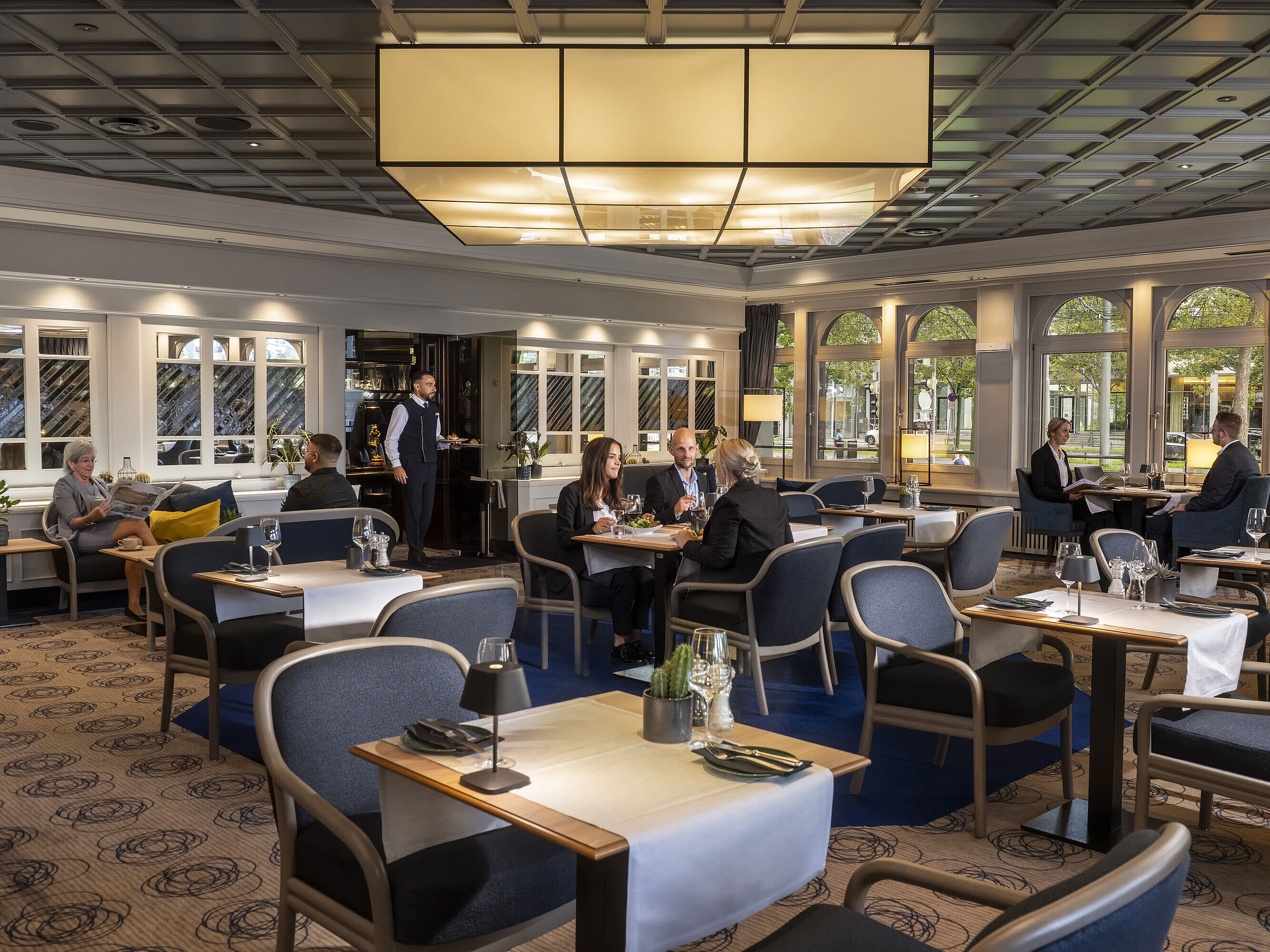 Elegant restaurant at Maritim Hotel Darmstadt with guests dining and relaxing. Modern decor and large windows with a view.