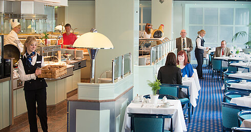 A waitress brings fresh bread rolls to the breakfast area in Maritim Hotel Magdeburg, while guests are seated at the table.