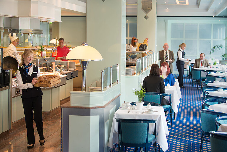 A waitress brings fresh bread rolls to the breakfast area in Maritim Hotel Magdeburg, while guests are seated at the table.
