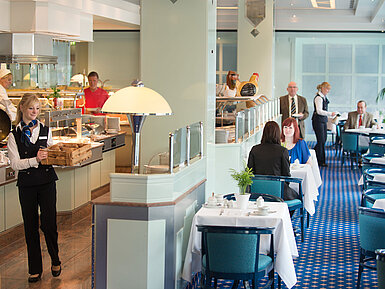A waitress brings fresh bread rolls to the breakfast area in Maritim Hotel Magdeburg, while guests are seated at the table.