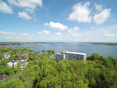 Aerial view of the Maritim Hotel Kiel, surrounded by green countryside and the Kiel Fjord, under a bright blue sky.