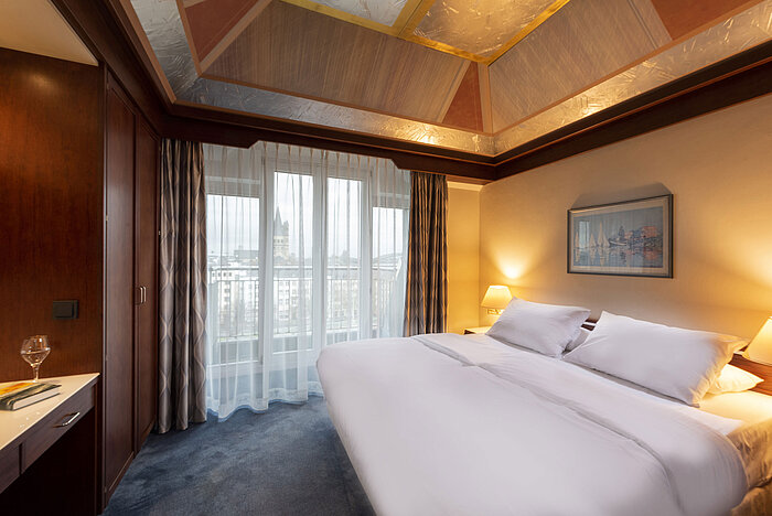 Junior Suite at Maritim Hotel Cologne with a large bed, desk, elegant ceiling, and panoramic view through large windows.