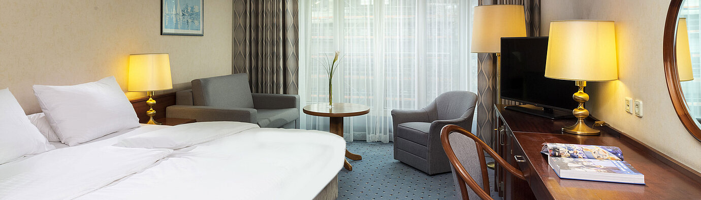 Bright family room at Maritim Hotel Cologne with double bed, desk, TV, armchairs, and large windows with curtains.