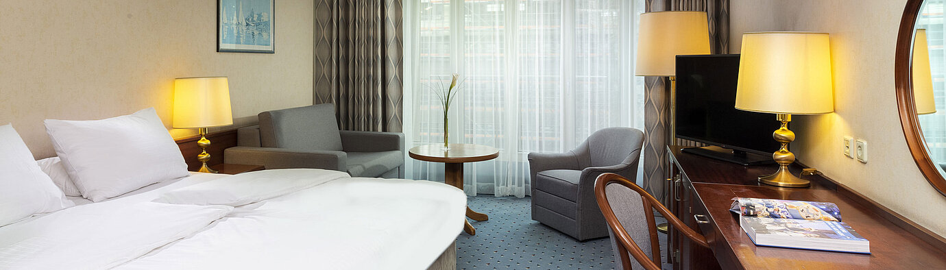 Bright family room at Maritim Hotel Cologne with double bed, desk, TV, armchairs, and large windows with curtains.