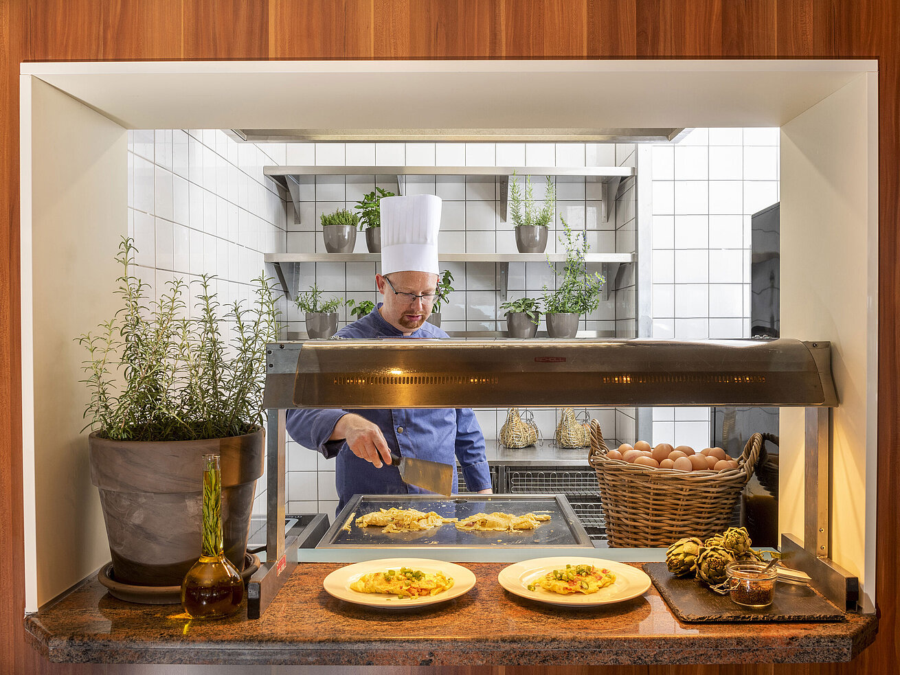 A chef prepares omelets at a cooking station surrounded by fresh herbs. Two plates of food are ready.