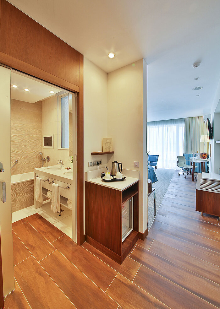 Bright hotel room with modern bathroom, tea station and view into the cosy living and sleeping area