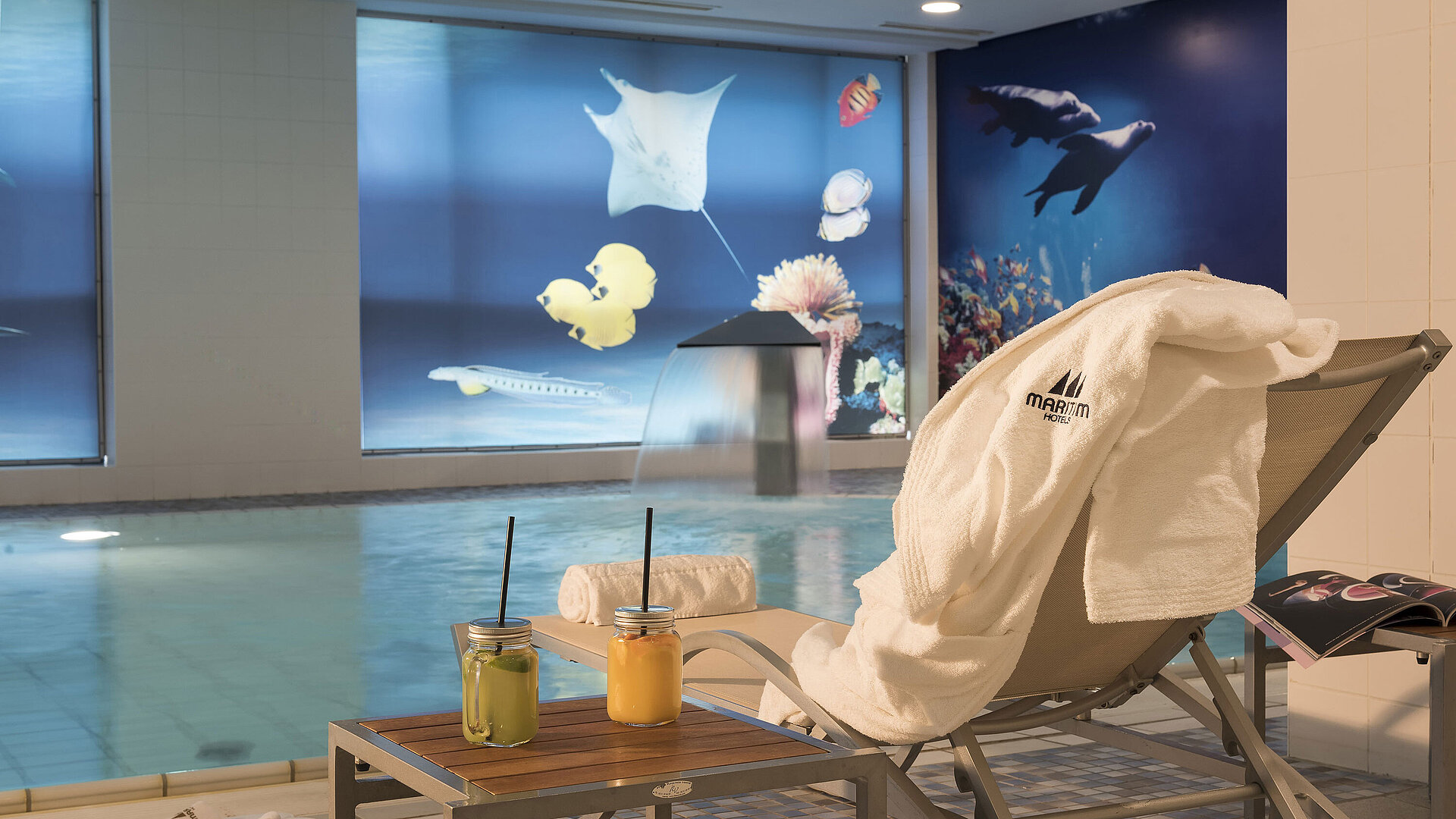 Relaxation area by the pool at the Maritim proArte Hotel Berlin with a lounger, towel, and fresh juices next to a waterfall.