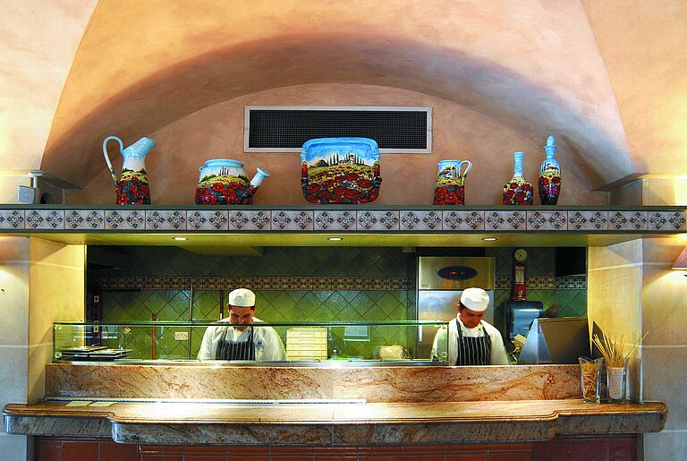 Open hotel kitchen with chefs preparing dishes behind a counter with Mediterranean decor