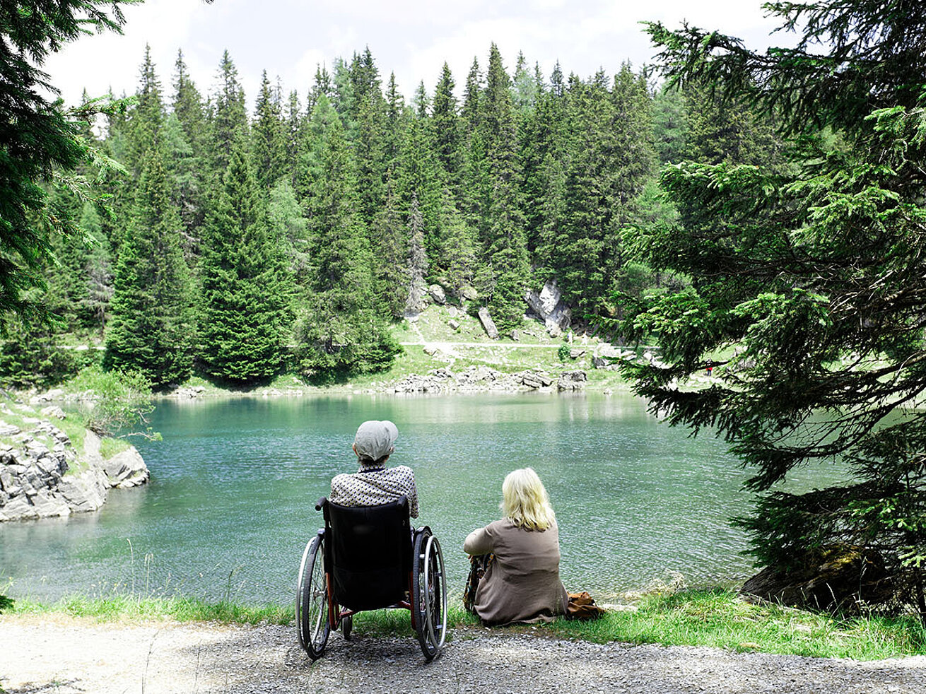 © Herby Meseritsch – AdobeStock.com Two people, one in a wheelchair, sitting by a lake and looking at the forest