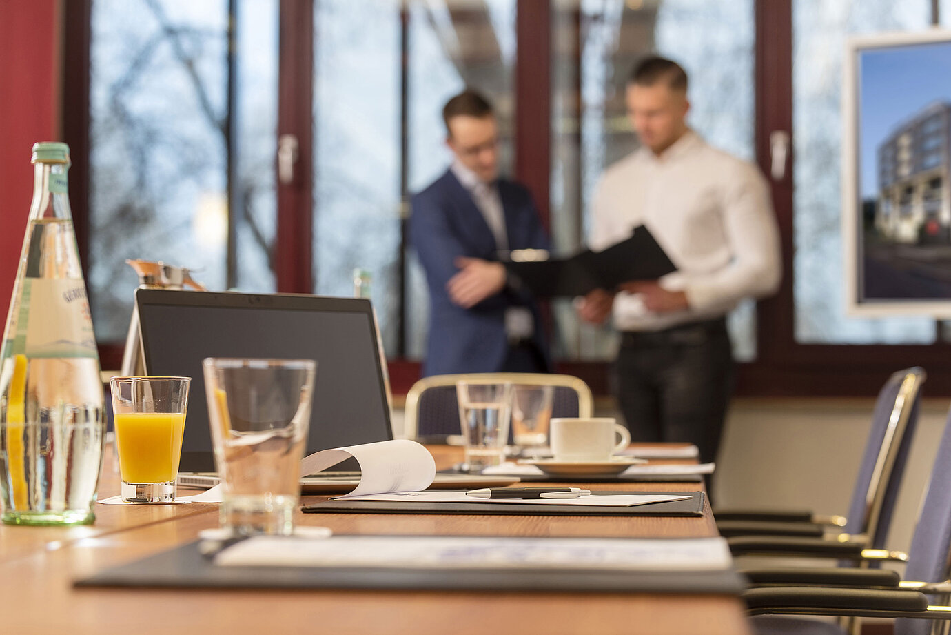 Conference room with a laptop, drinks, and documents, two businessmen in the background discussing papers.