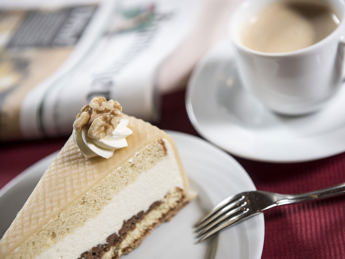 Slice of cream cake on a plate with a fork, next to a cup of coffee and a newspaper.