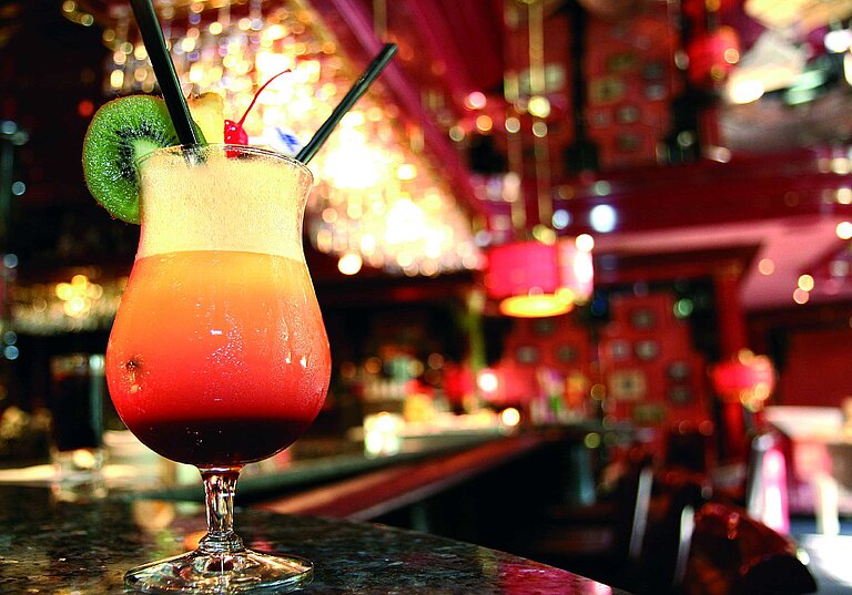 Colourful cocktail with lime and cherry on the bar counter, warm-lit hotel bar in the background.