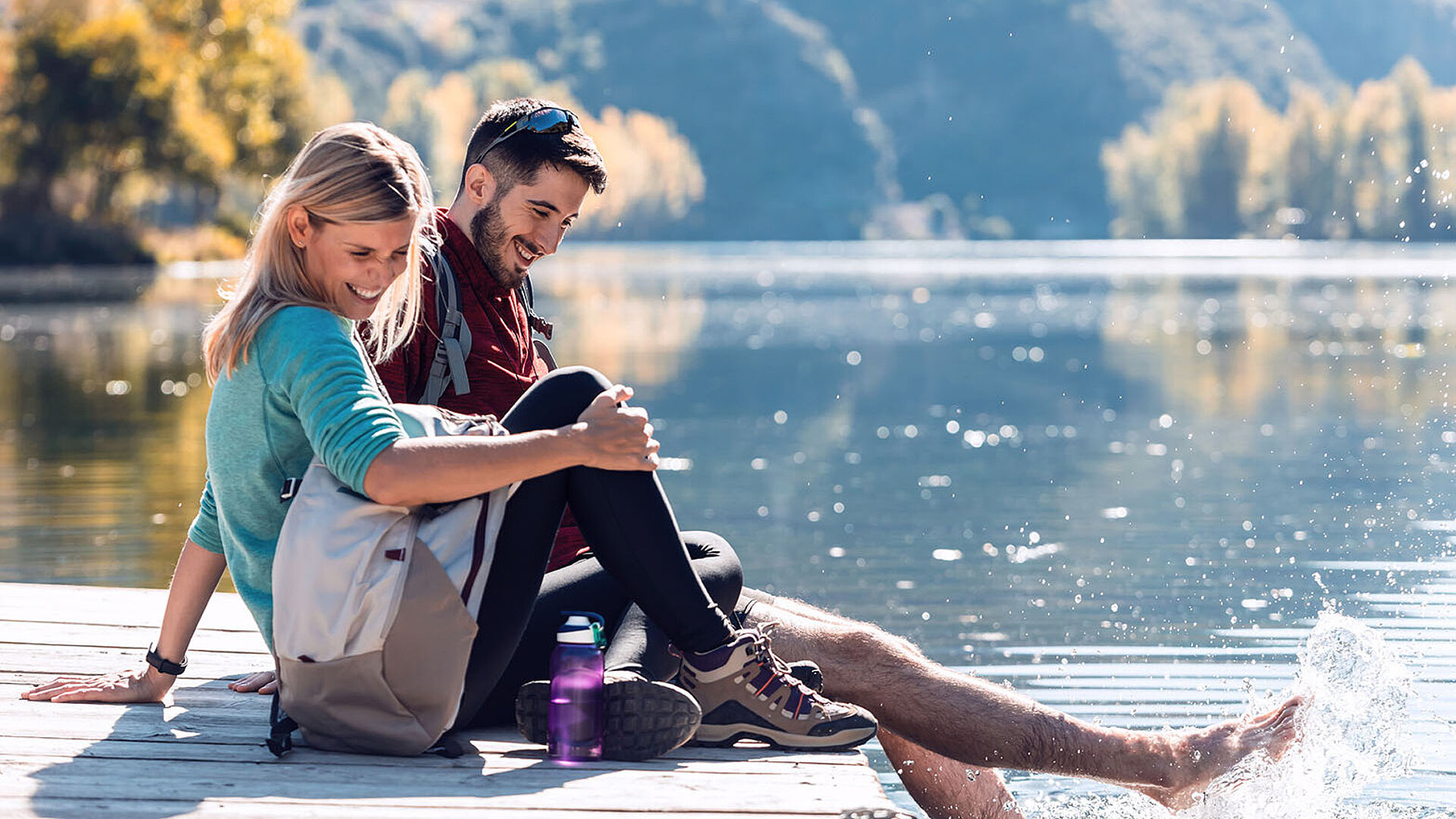 Smiling couple sitting on a jetty with backpacks, splashing feet in the clear water of a mountain lake