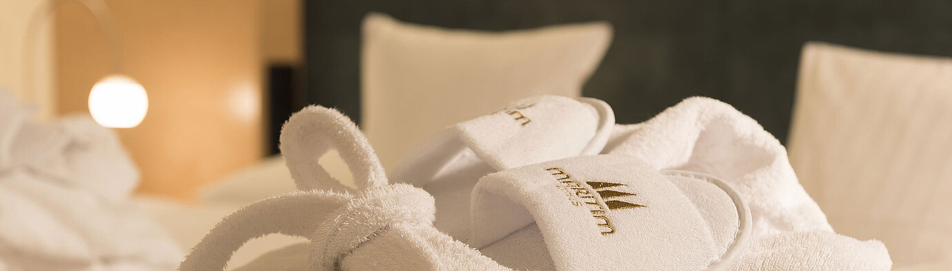 White bathrobe and slippers with Maritim logo placed on the bed for relaxing wellness moments in the hotel room