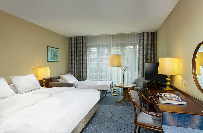 Bright family room at Maritim Hotel Cologne with double bed, desk, TV, armchairs, and large windows with curtains.