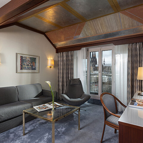 Junior Suite at Maritim Hotel Cologne with a view of Cologne Cathedral, stylish sofa, chair, and desk with reading material.