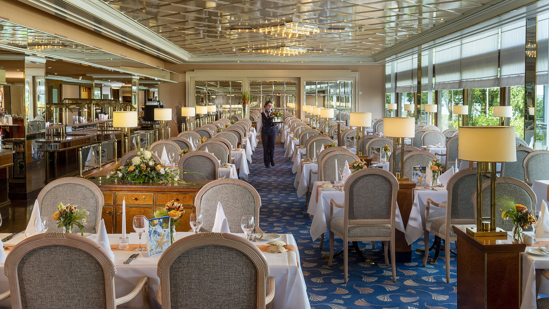 Large, elegant restaurant with long set tables, chandeliers, and floral arrangements for fine dining.