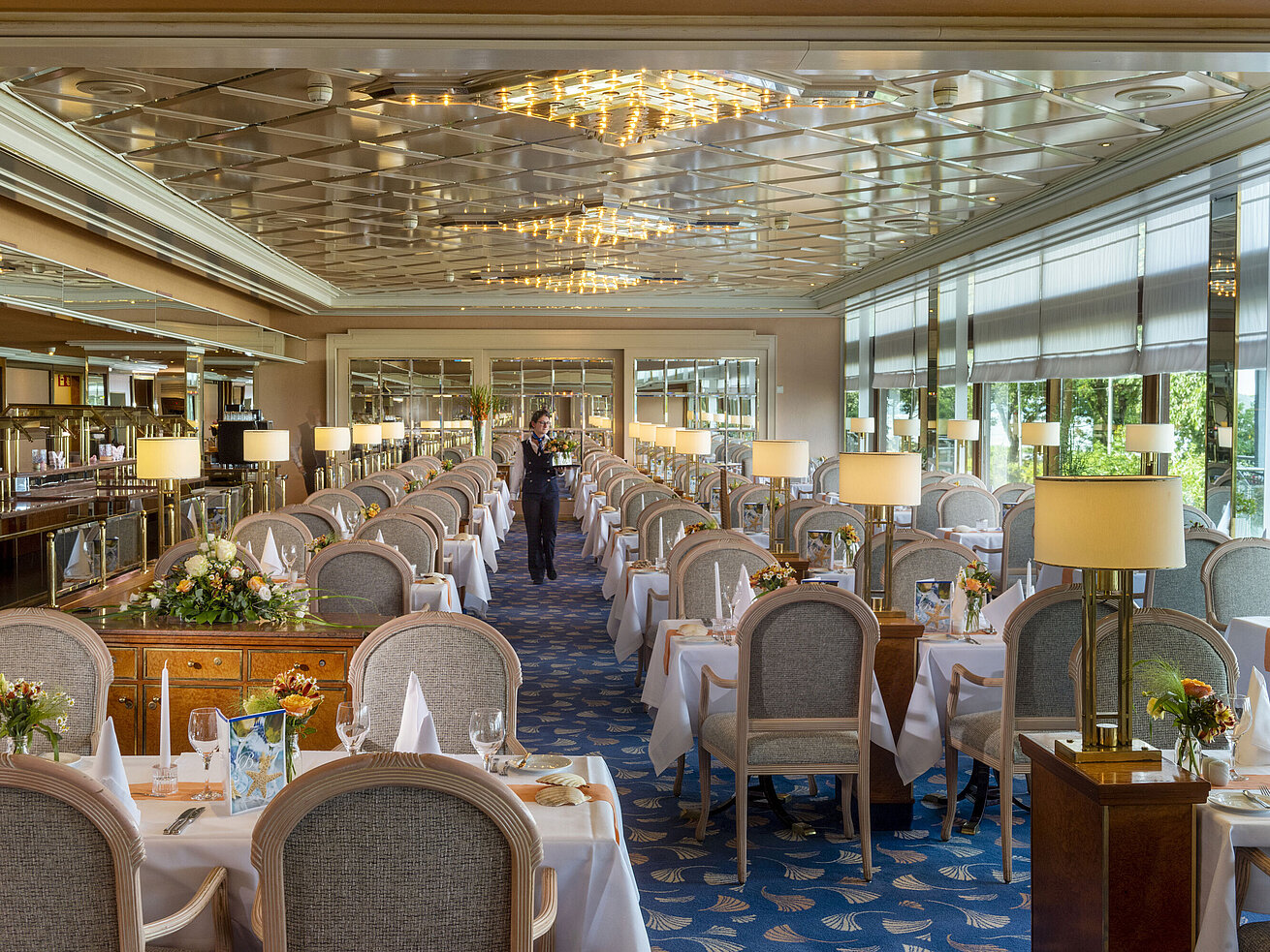 Large, elegant restaurant with long set tables, chandeliers, and floral arrangements for fine dining.