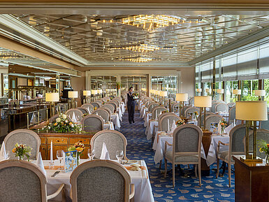 Large, elegant restaurant with long set tables, chandeliers, and floral arrangements for fine dining.