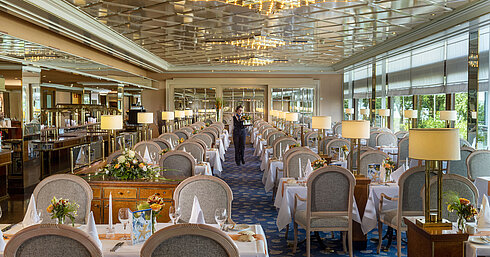 Large, elegant restaurant with long set tables, chandeliers, and floral arrangements for fine dining.