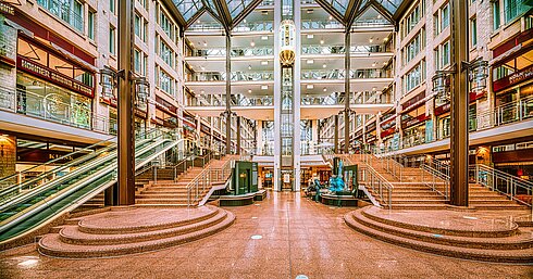 The spacious lobby of the Maritim Hotel Cologne with stairs, escalators, glass roof, and shops on multiple levels.