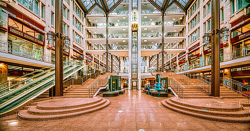 The spacious lobby of the Maritim Hotel Cologne with stairs, escalators, glass roof, and shops on multiple levels.
