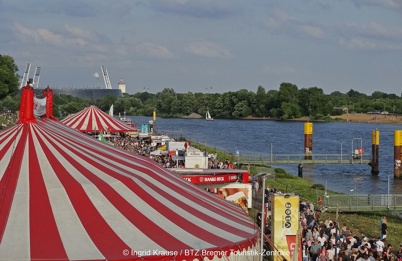 Crowds and colorful tents at the Weser River during an event in Bremen, with a view of the stadium and river