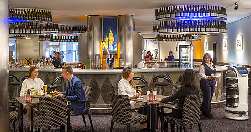 Guests dining at the bar of the Maritim proArte Hotel Berlin, surrounded by modern facilities and a service robot.